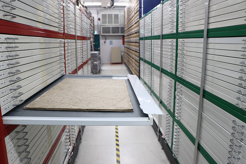 A drawer containing a pile of large map sheets. The corridor is clean and brightly lit. The drawers are about two metres tall and line both sides.